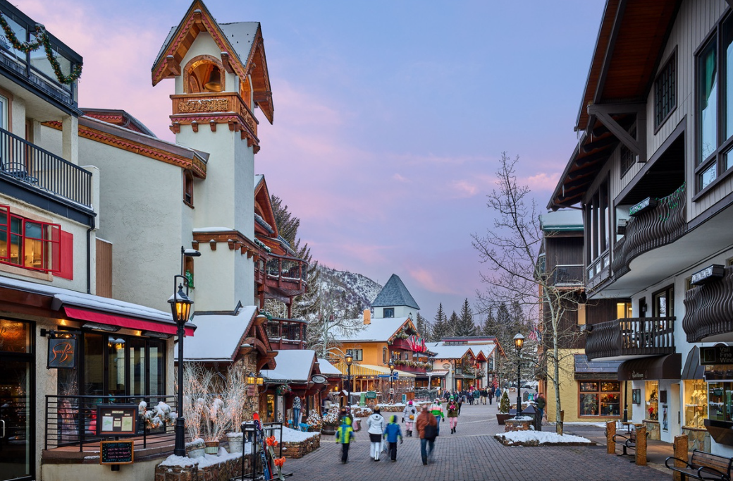 People walk along a snowy alpine village street lined with colorful buildings and balconies beneath a pink evening sky.