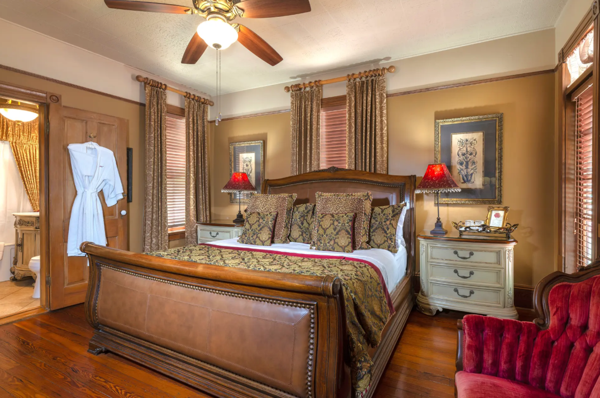 Traditional bedroom with wooden sleigh bed, patterned pillows, red lamps, ceiling fan, and robe by the bathroom door.