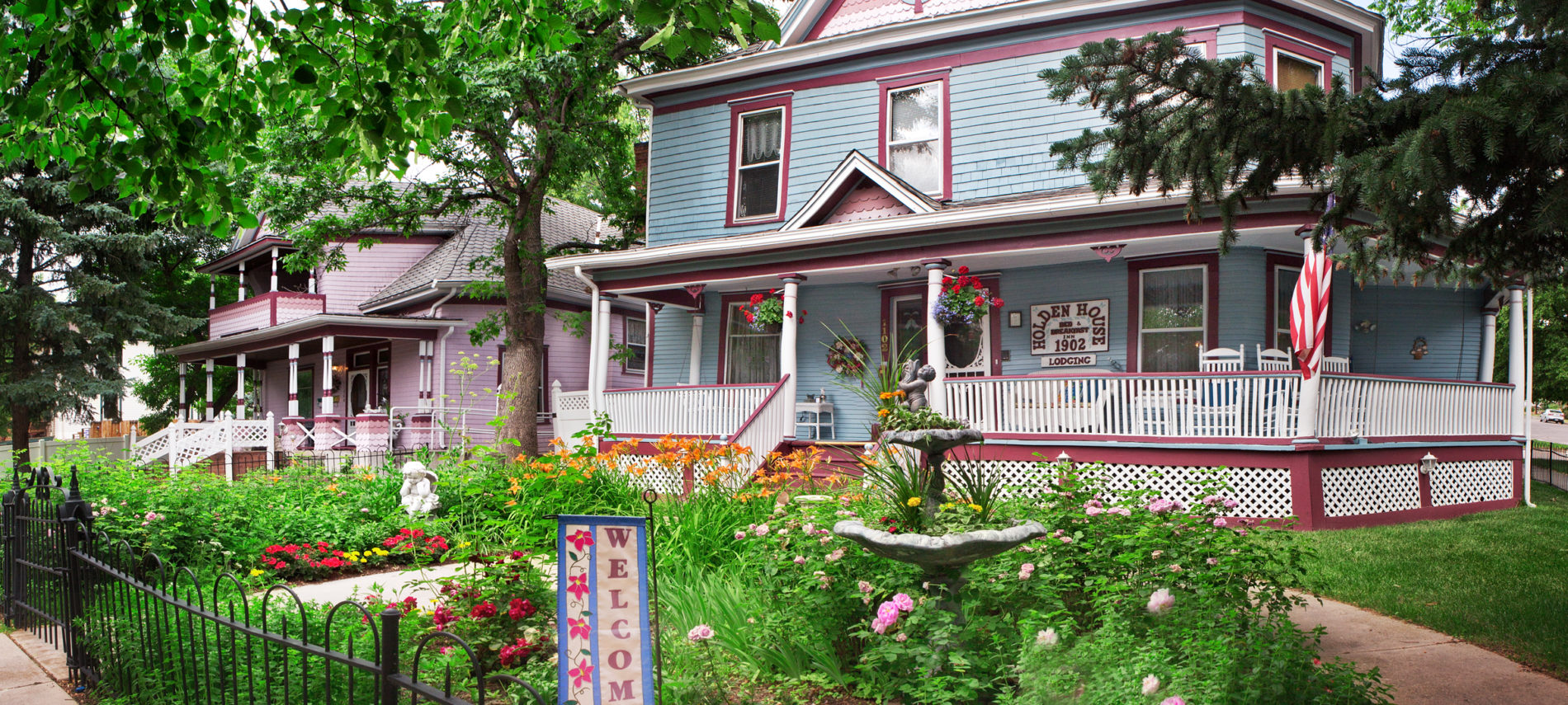 Two Victorian-style houses with wraparound porches sit amid lush gardens, flags, and a front-yard "Welcome" sign.