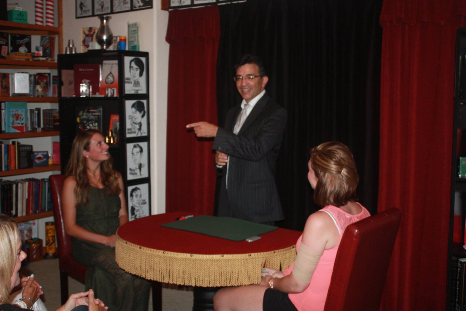 A man in a suit gestures while two women sit at a round table with a red cloth in a room lined with books and framed photos.