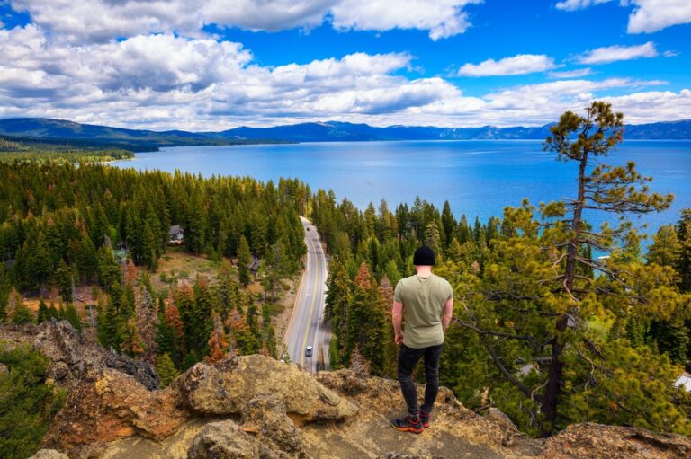 A person stands on a rocky outcrop, overlooking a forest, winding road, and large lake beneath a partly cloudy sky.