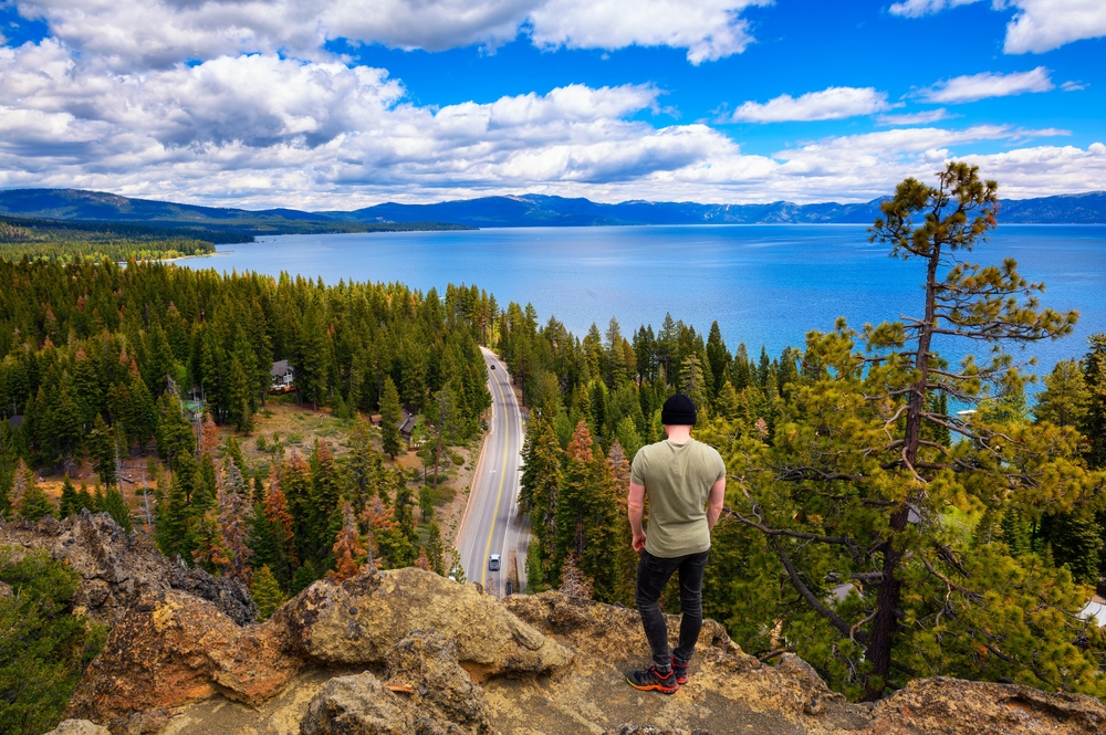 A person stands on a rocky outcrop, overlooking a forest, winding road, and large lake beneath a partly cloudy sky.