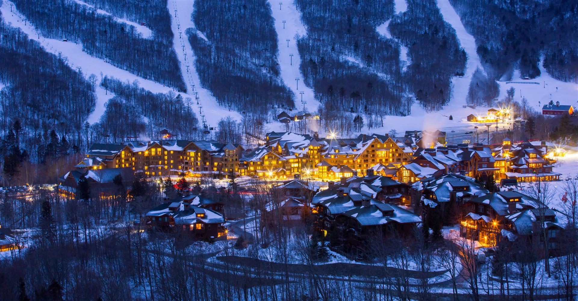 At night, a ski resort's lit buildings sit at the snowy slope base with ski lifts and trees surrounding the scene.
