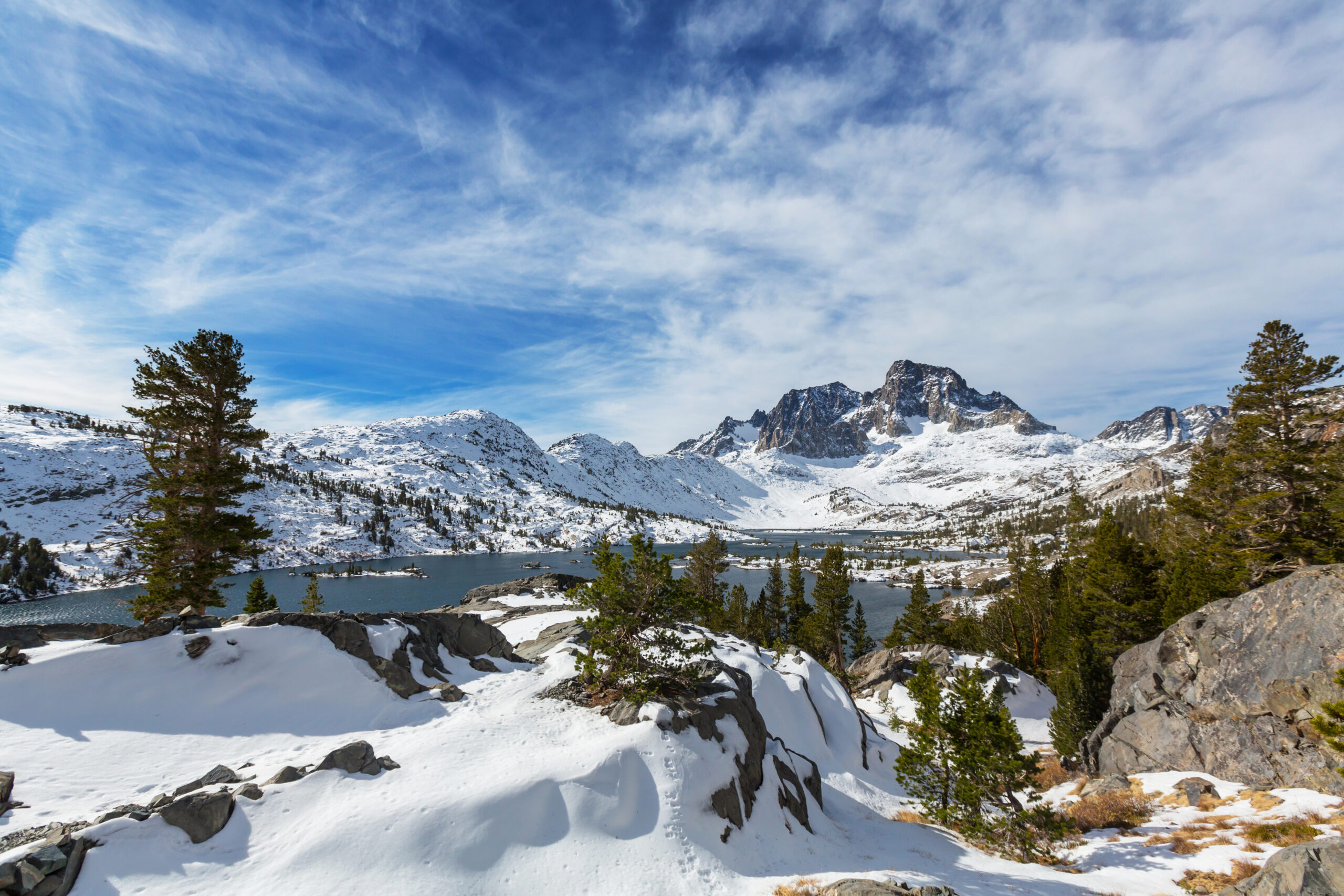 Snowy mountains and pines encircle a lake beneath partly cloudy skies, with rocky ground in the foreground.