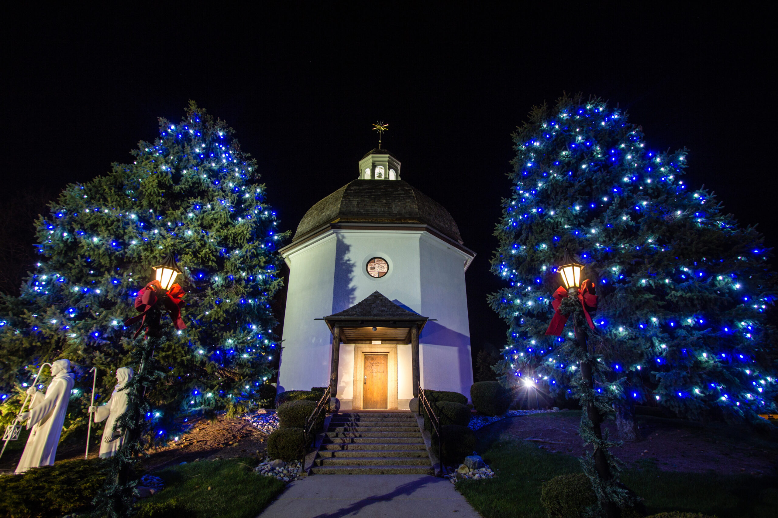A small chapel sits at night between blue-lit trees, with street lamps adorned in red bows and white statues to the left.