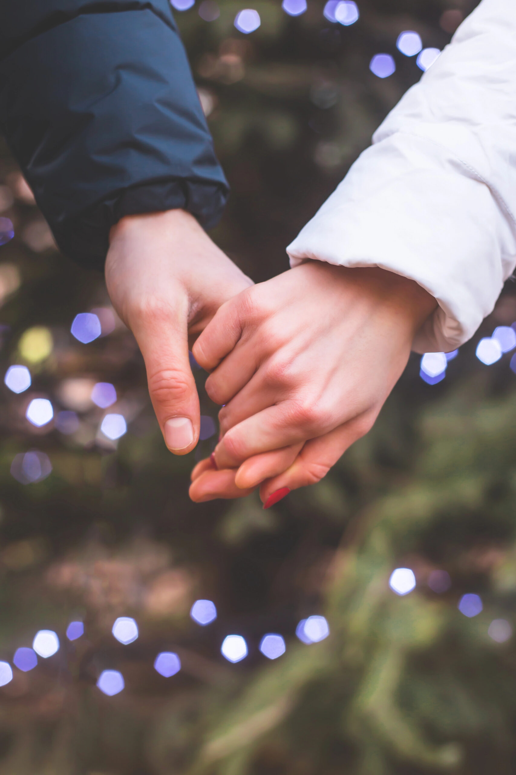 Two people holding hands, one in dark and one in white sleeves, with blurred lights and greenery creating a romantic holiday scene.