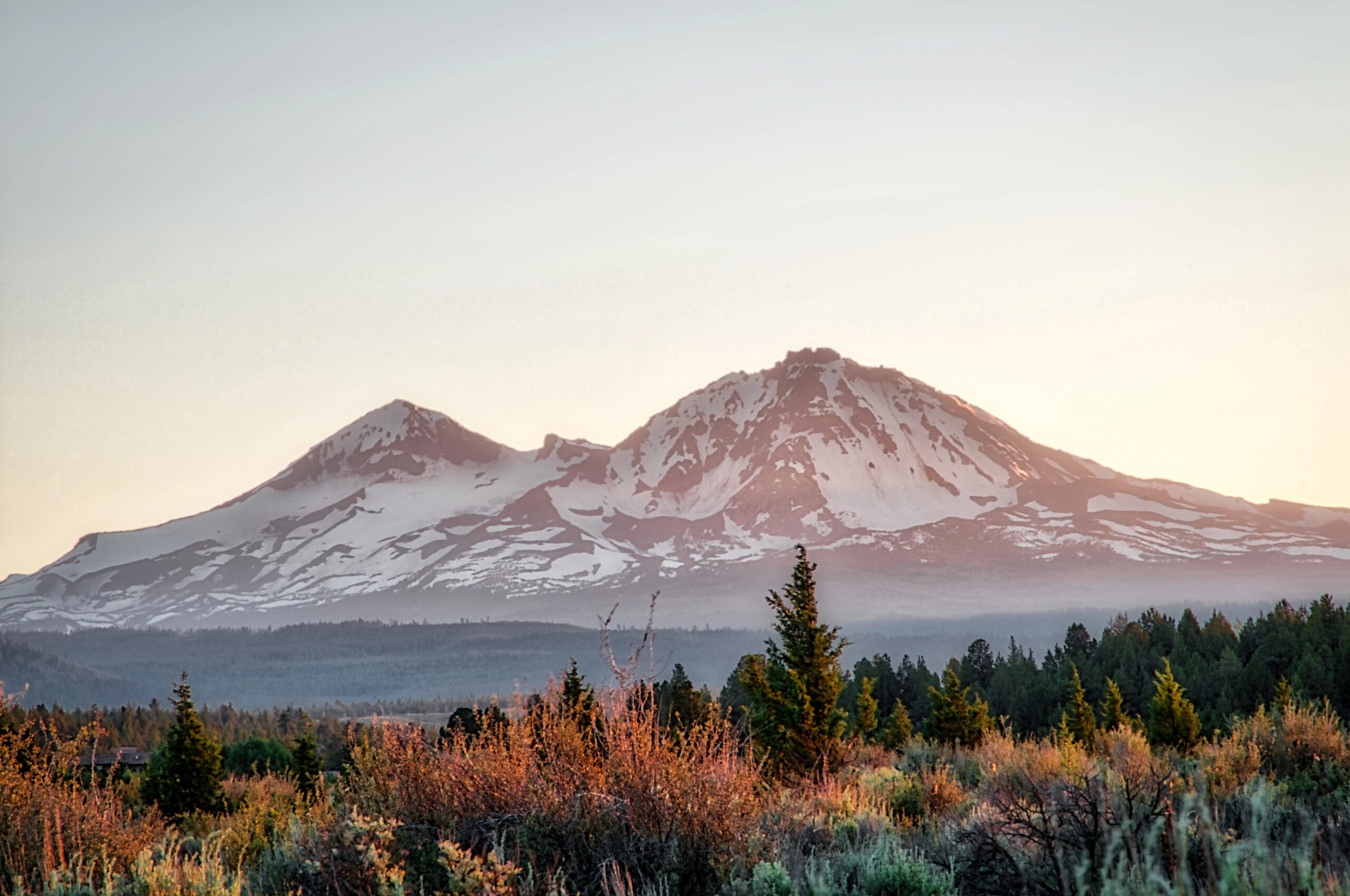 Snow-capped peaks tower over a sunlit forest at sunrise, creating a perfect Christmas getaway scene for couples.