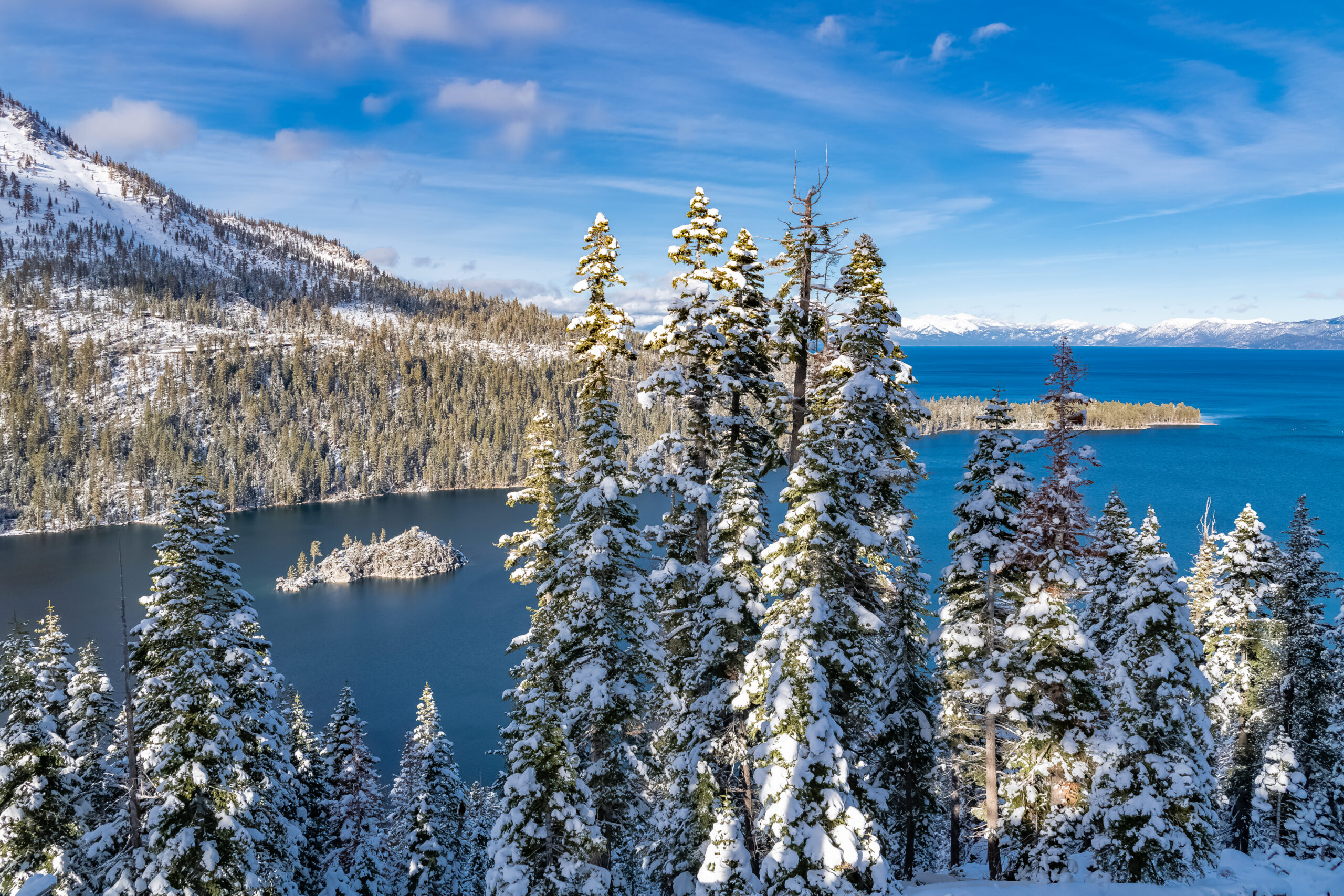 Snow-covered pines overlook a blue lake with an island and forested mountains beneath a partly cloudy sky.