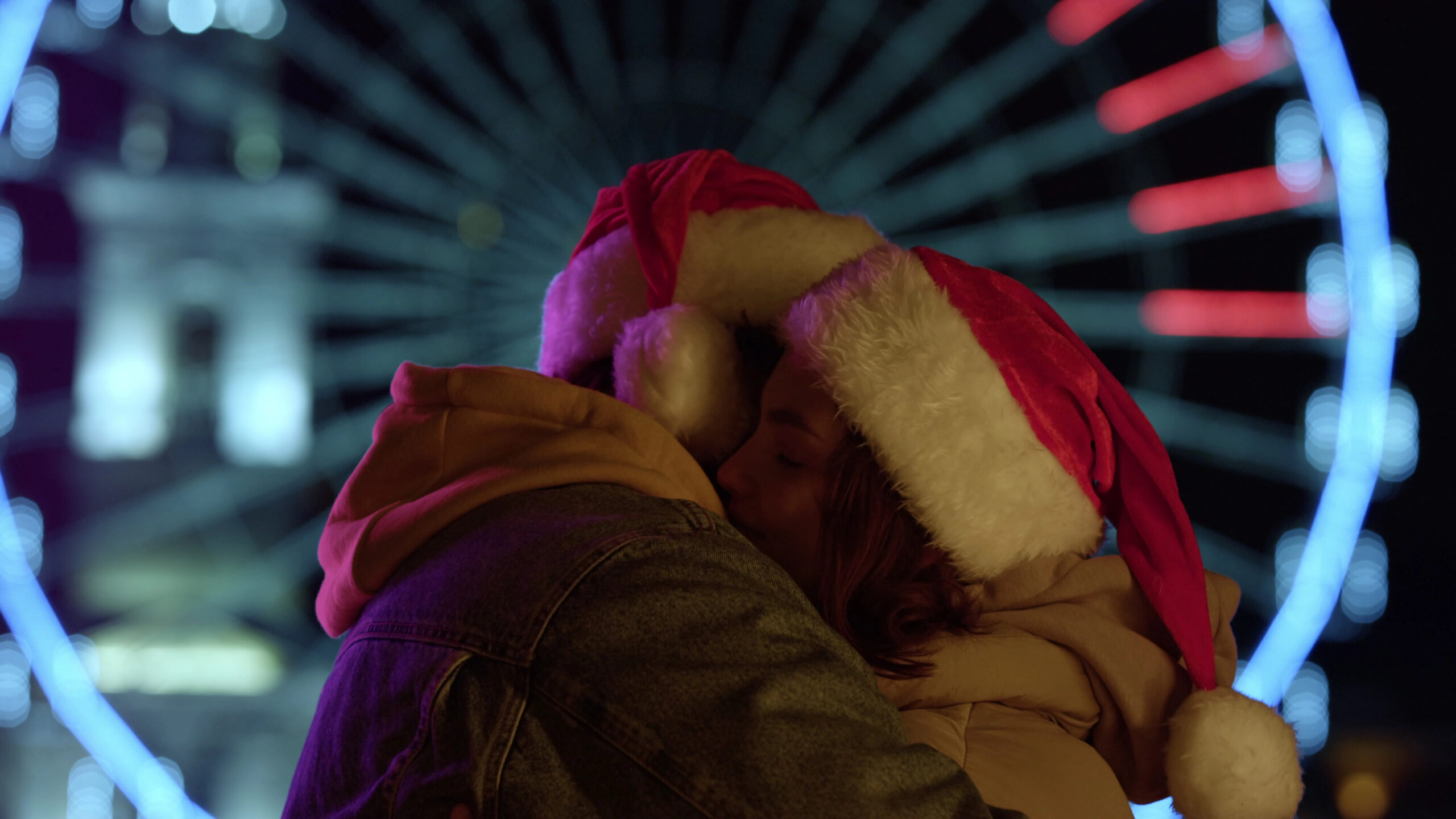 Two people in Santa hats embrace at night before a glowing Ferris wheel, evoking the magic of Christmas couple vacations.