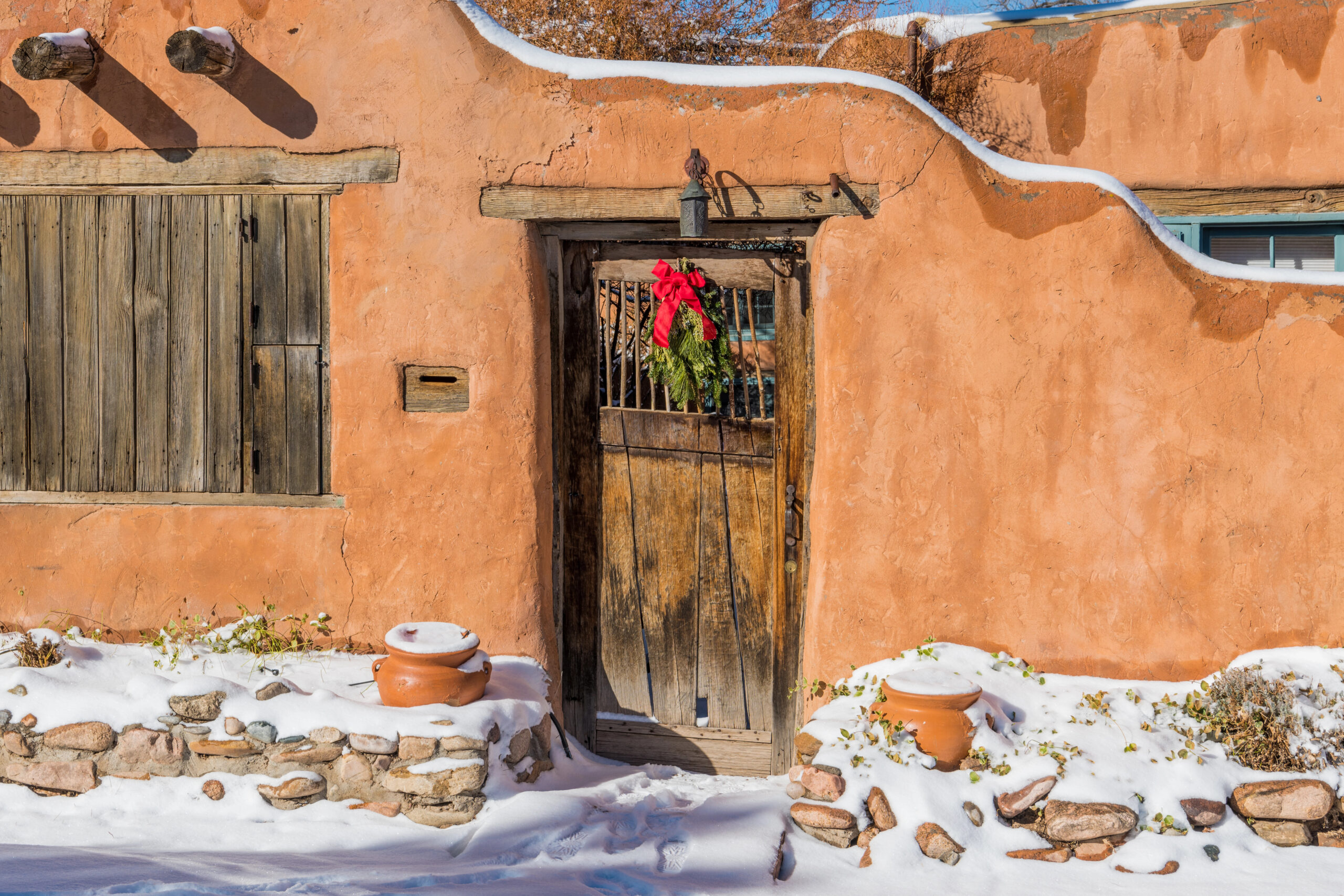 A rustic adobe wall and wooden door with a holiday wreath and bow, snow on the ground, and potted plants nearby.