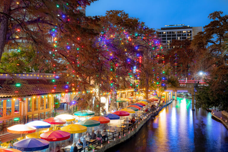 Colorful umbrellas and string lights brighten the San Antonio River Walk at dusk, reflecting magically in the water.