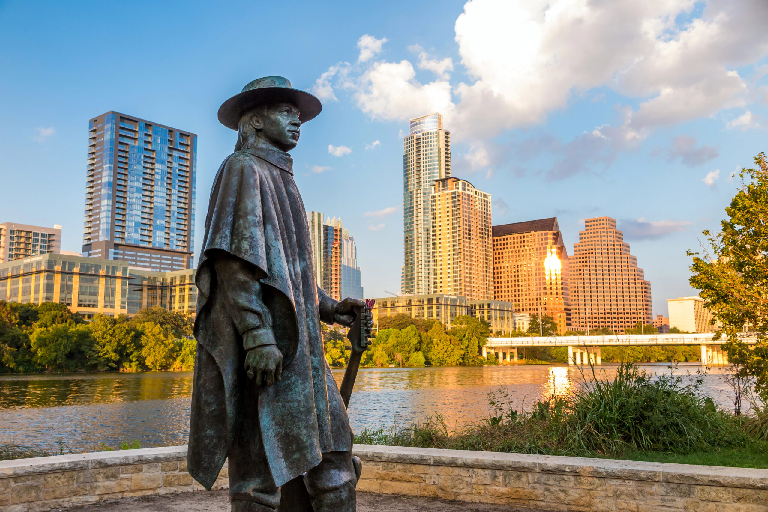 Bronze statue holding a guitar stands by a river, backed by city buildings, trees, and partly cloudy skies.