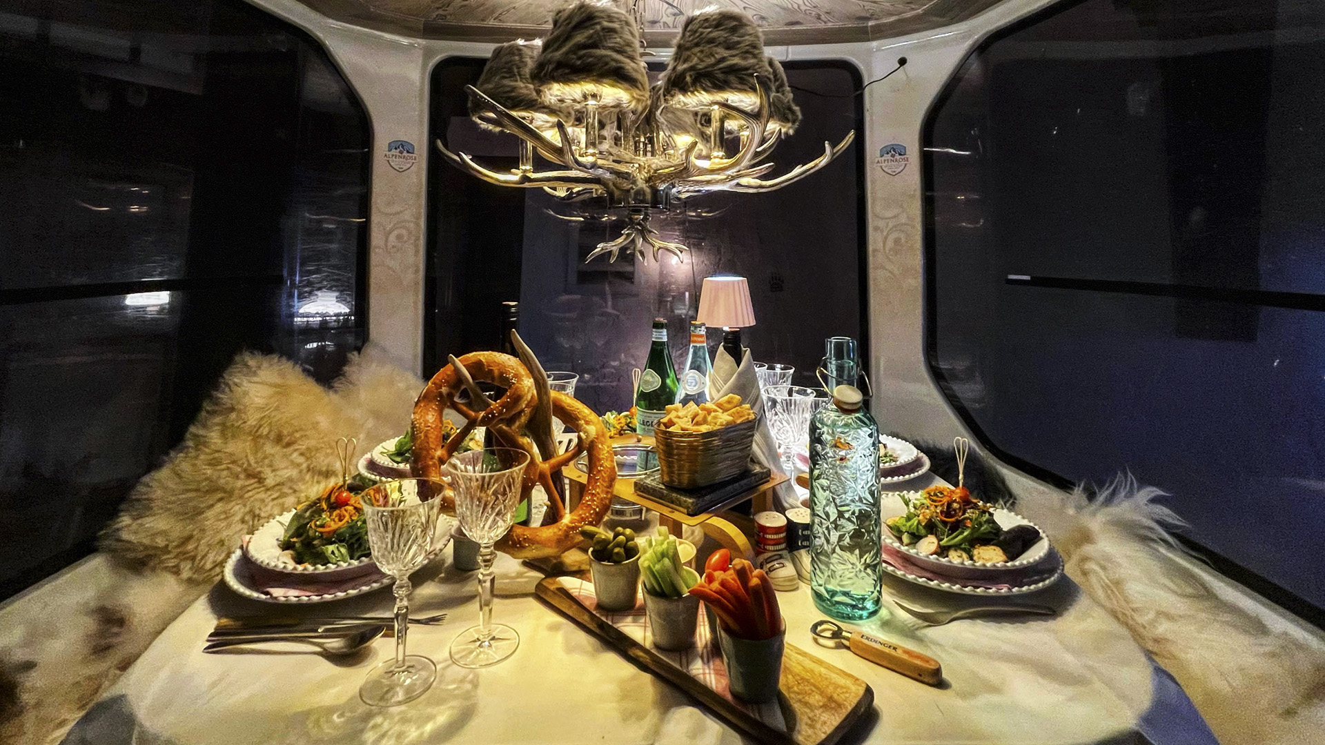 A cozy gondola dining table for two with salads, snacks, wine glasses, and fur seats beneath a modern chandelier.