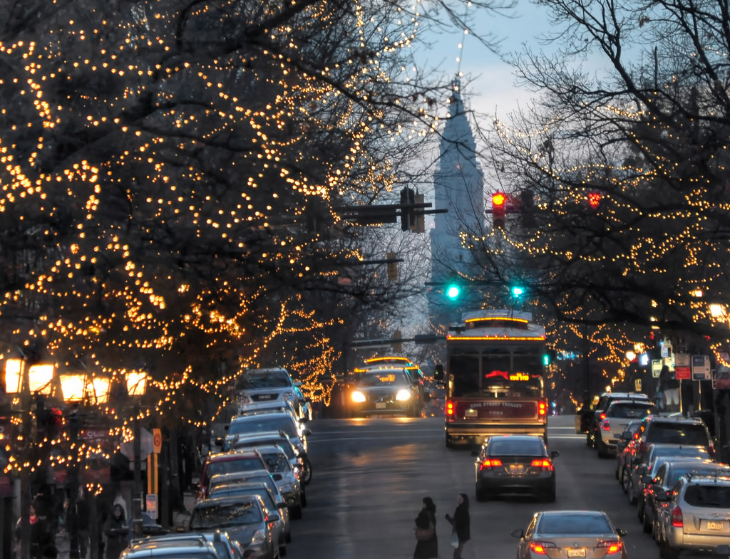 At dusk, cars line both sides of a city street with string-lit trees and distant traffic lights.