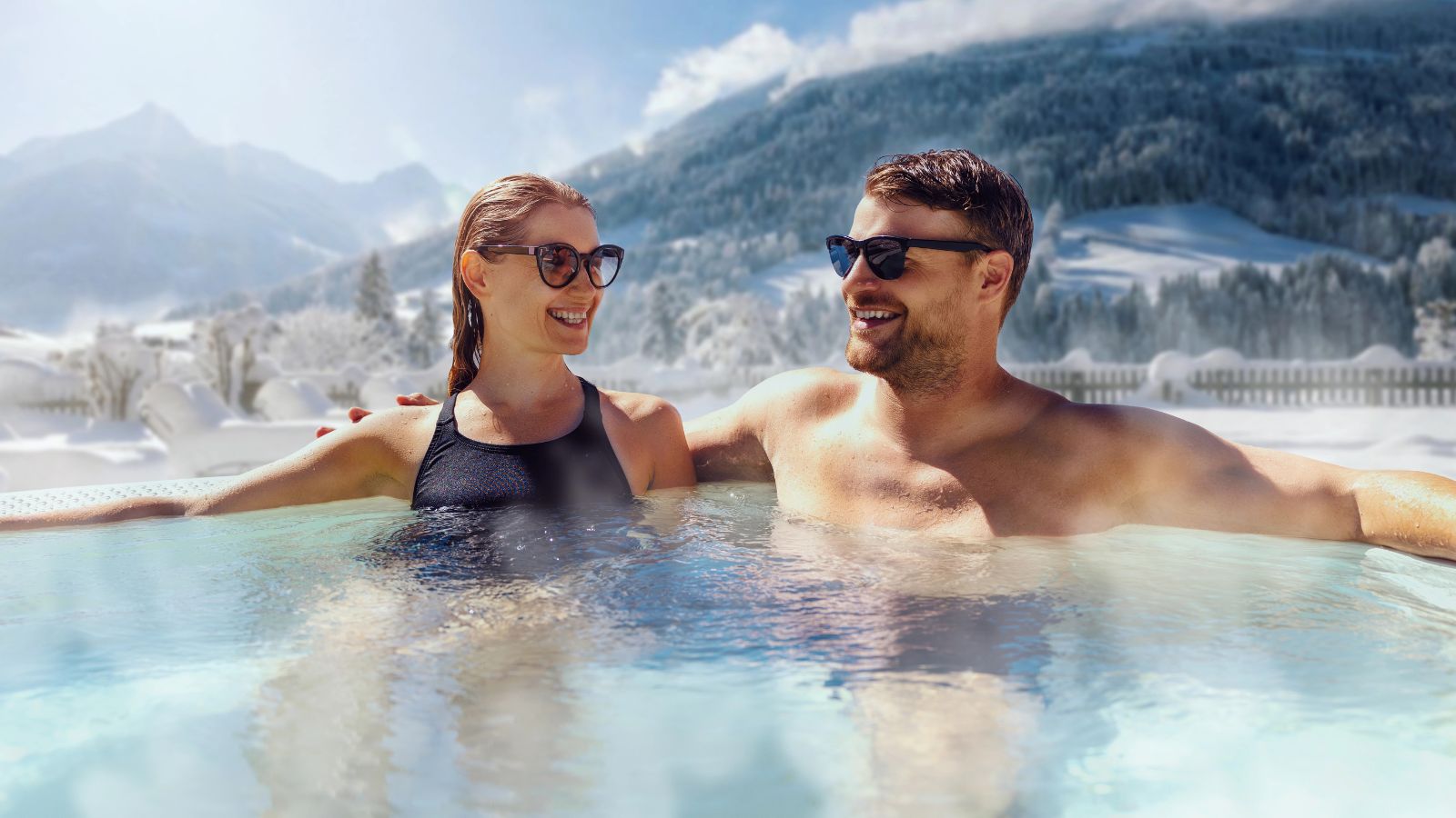 A man and woman in swimsuits smile together in an outdoor hot tub with snowy mountains and trees behind them.