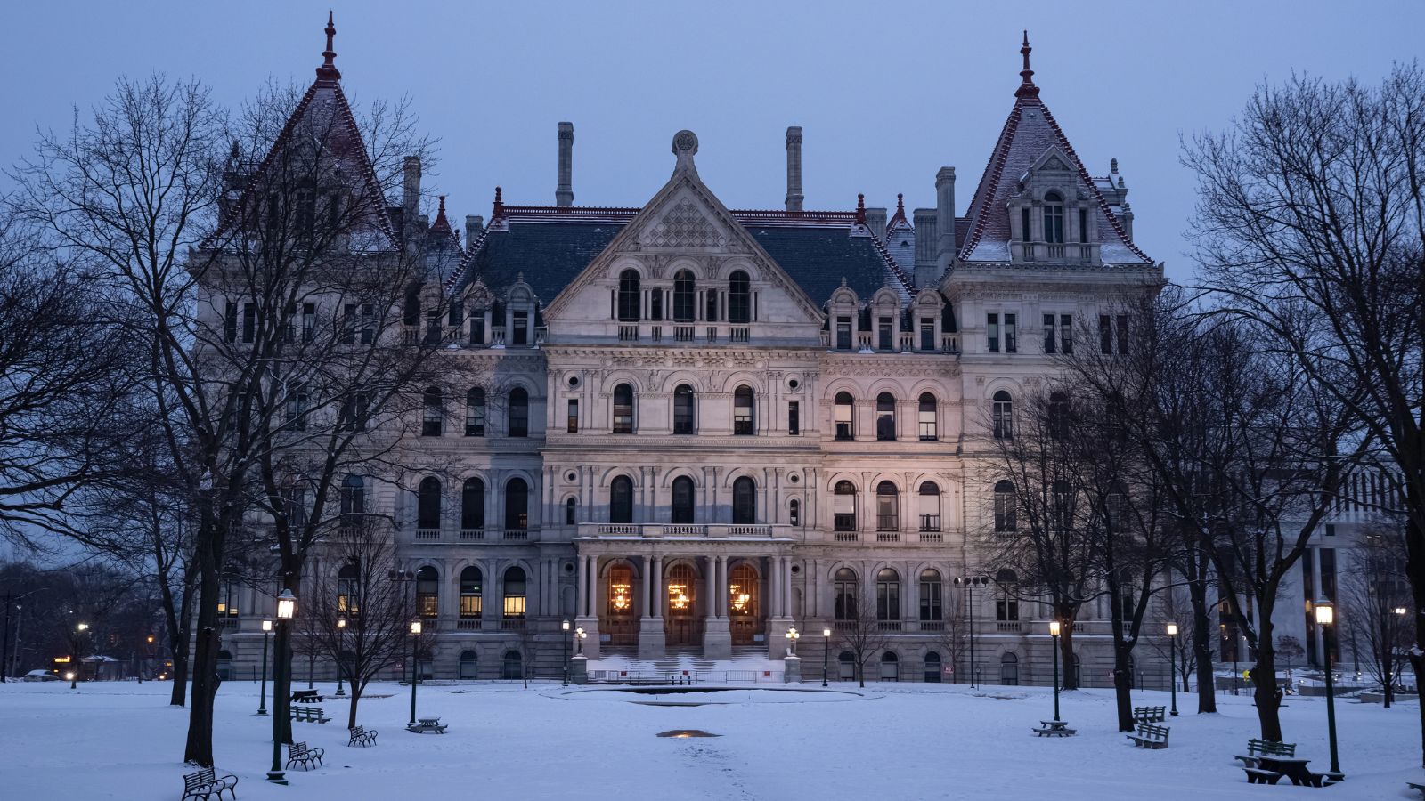 A gothic stone government building glows at dusk amid bare trees and snowy ground.