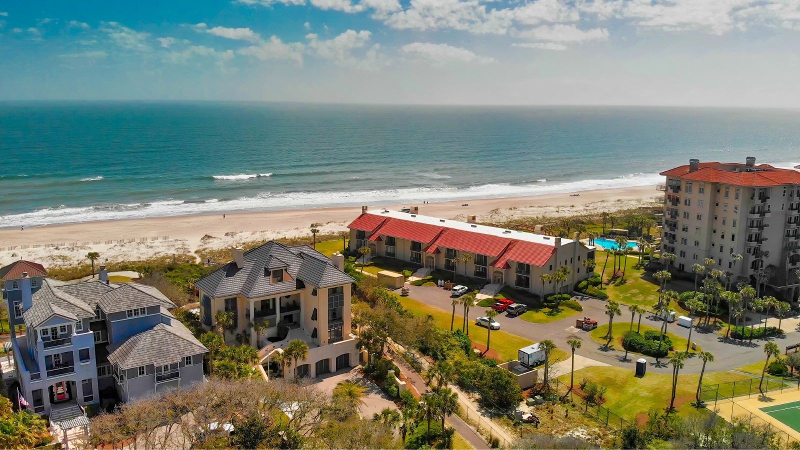 Aerial view of beachfront buildings along a sandy coast with the ocean in the background under partly cloudy skies.
