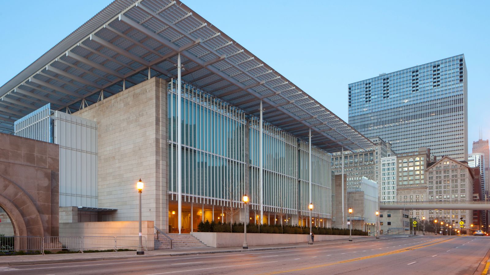 Modern building with glass facade and metal roof on an empty city street, skyscrapers visible in the background.