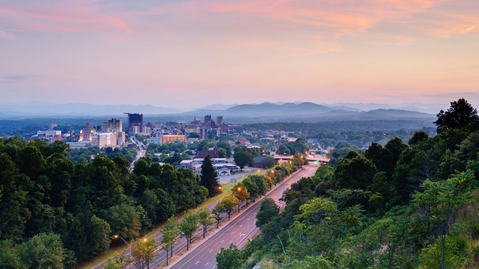 City skyline with trees, a highway in front, and mountains behind, all at sunset.