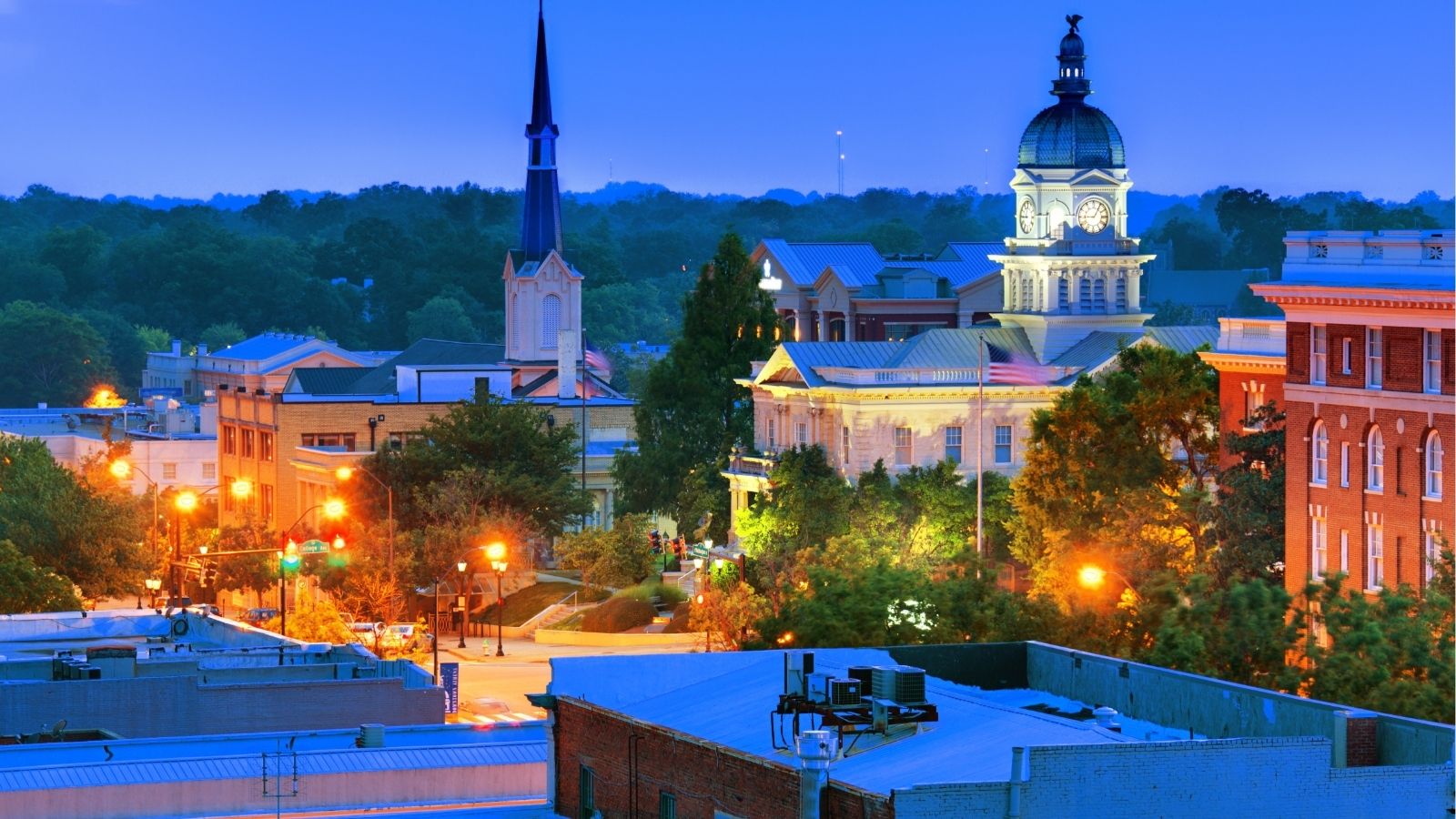 Evening downtown scene in a small city with historic buildings, a clock tower, church steeple, and glowing streetlights.