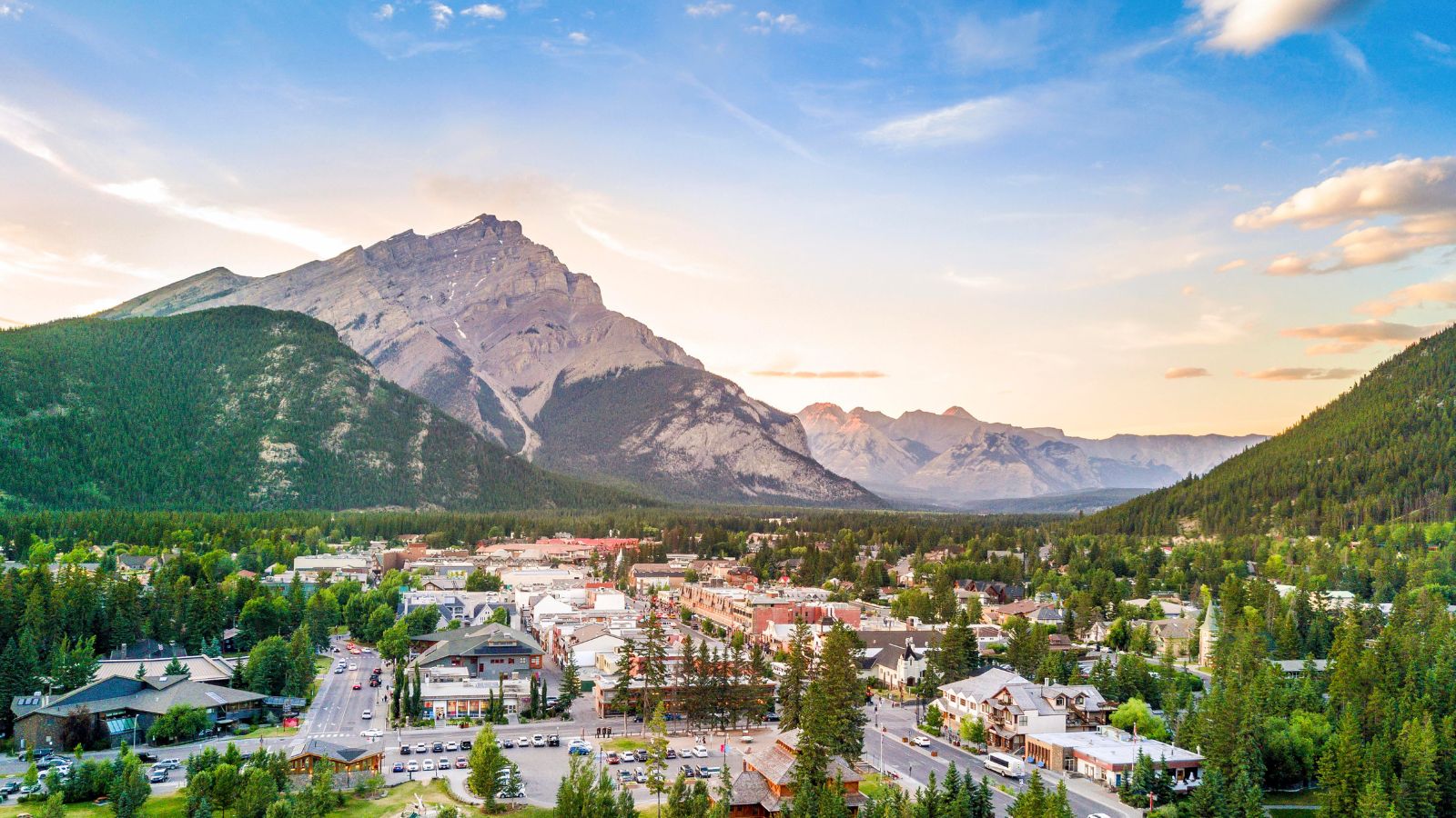 Aerial view of a small town bordered by pine forests and mountains beneath a partly cloudy sunset sky.