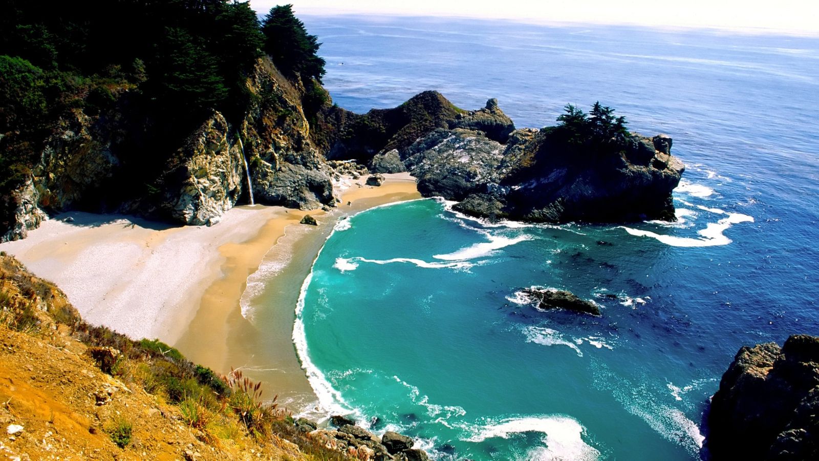 Secluded cove with turquoise water, sandy beach, rocky cliffs, and dense green trees under a clear sky.