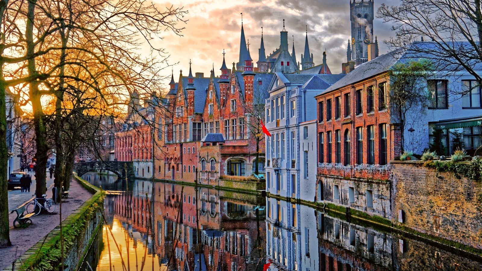 Colorful historic buildings and trees reflect in a Bruges, Belgium canal at sunset.
