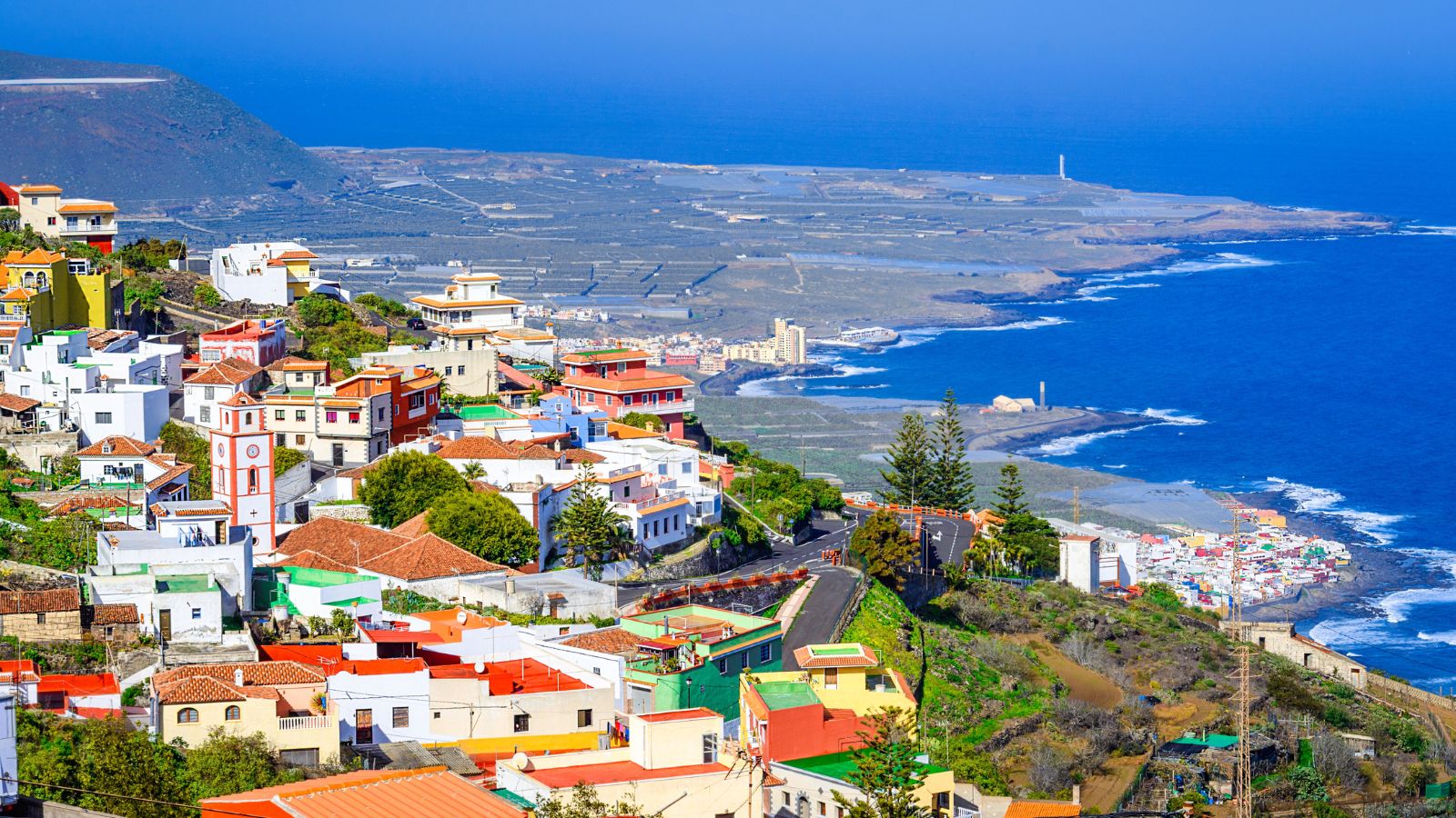 Colorful hillside houses overlook the ocean, with fields and buildings extending to the horizon beneath a clear sky.