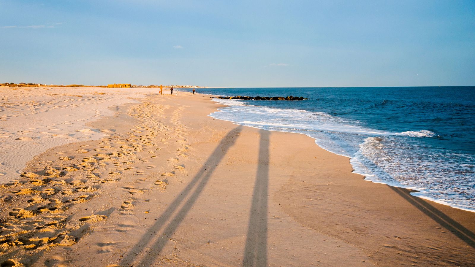 Footprints and long shadows stretch across a sandy beach at sunset, with gentle waves and distant walkers visible.