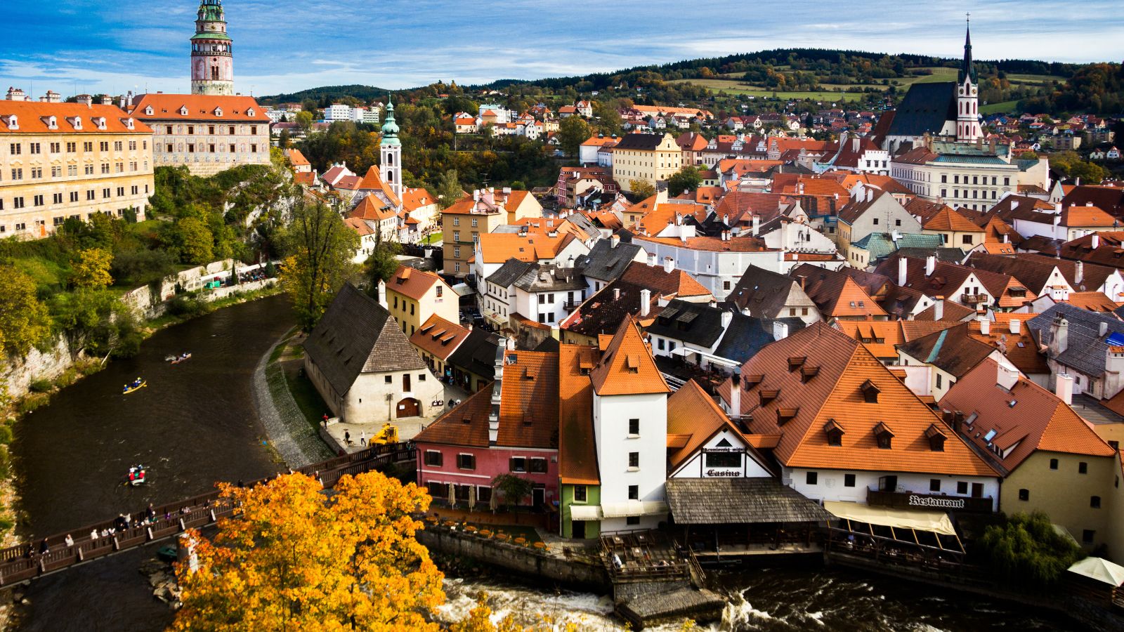 Panoramic view of a European town with red roofs, river kayakers, a stone bridge, and church spire beneath partly cloudy skies.