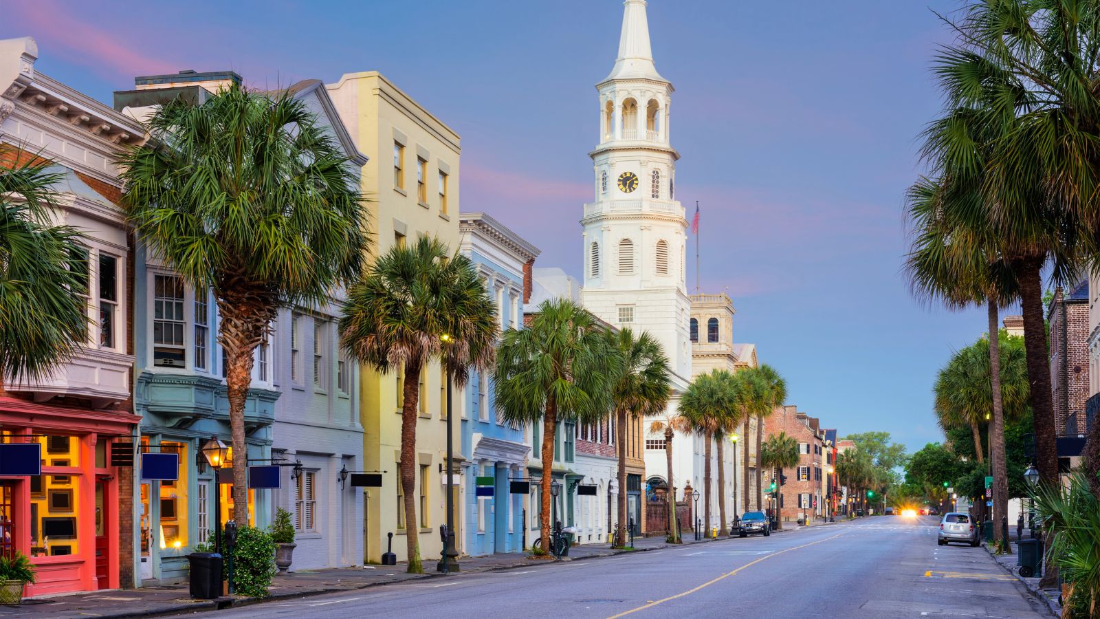 Colorful buildings and palm trees line a street, with a white church and clock tower in the background under a pastel sky.