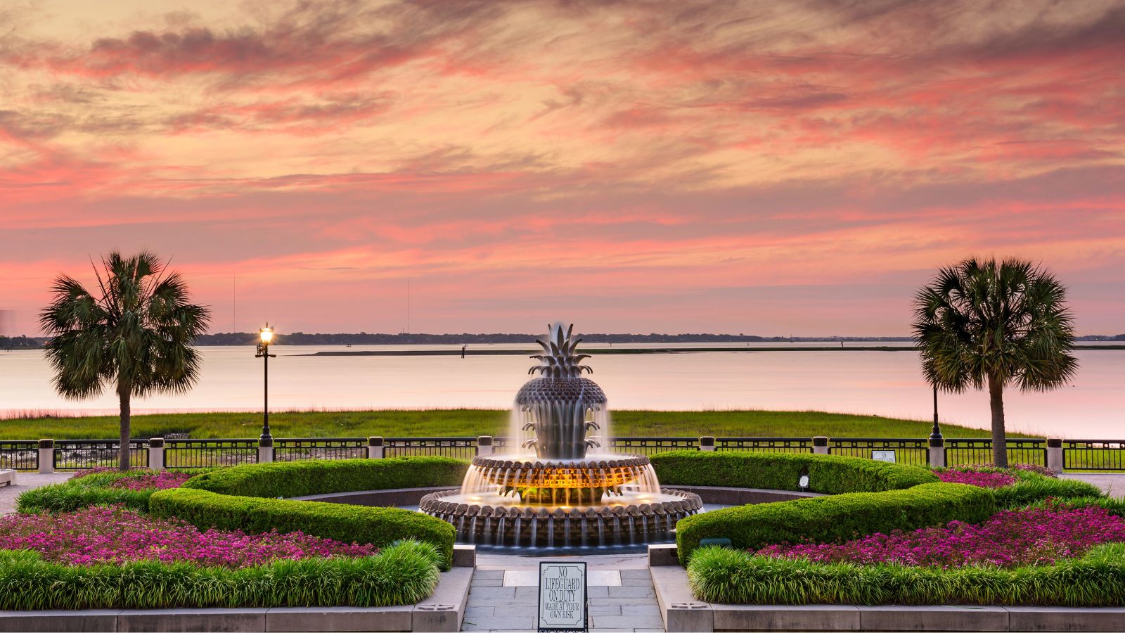 A pineapple-shaped fountain in landscaped gardens with palm trees overlooks a calm waterfront at sunset under a colorful sky.