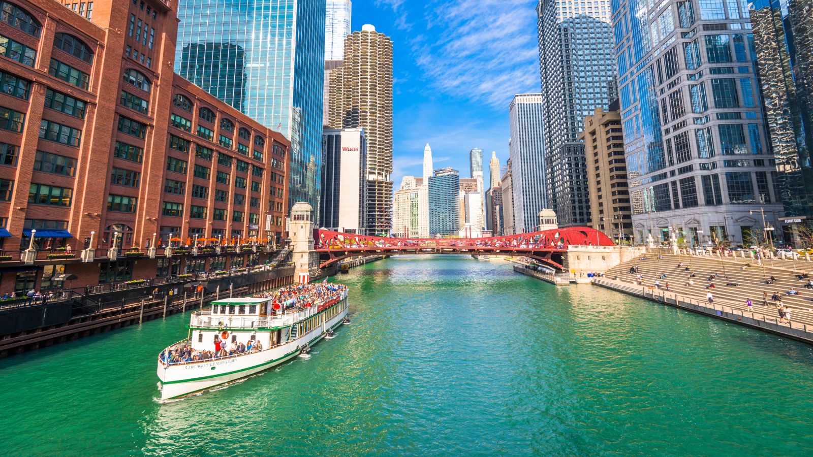 A tour boat cruises the Chicago River past tall modern and historic buildings, with a red bridge and blue sky behind.
