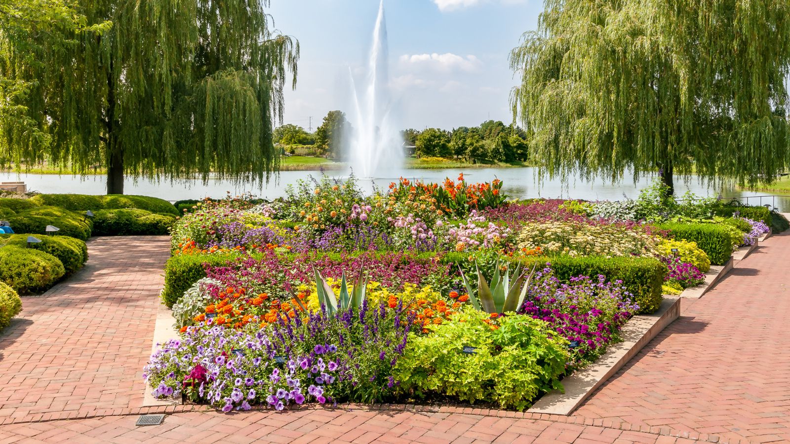 A vibrant flower garden with brick paths, two willow trees, and a lakefront fountain in the background.