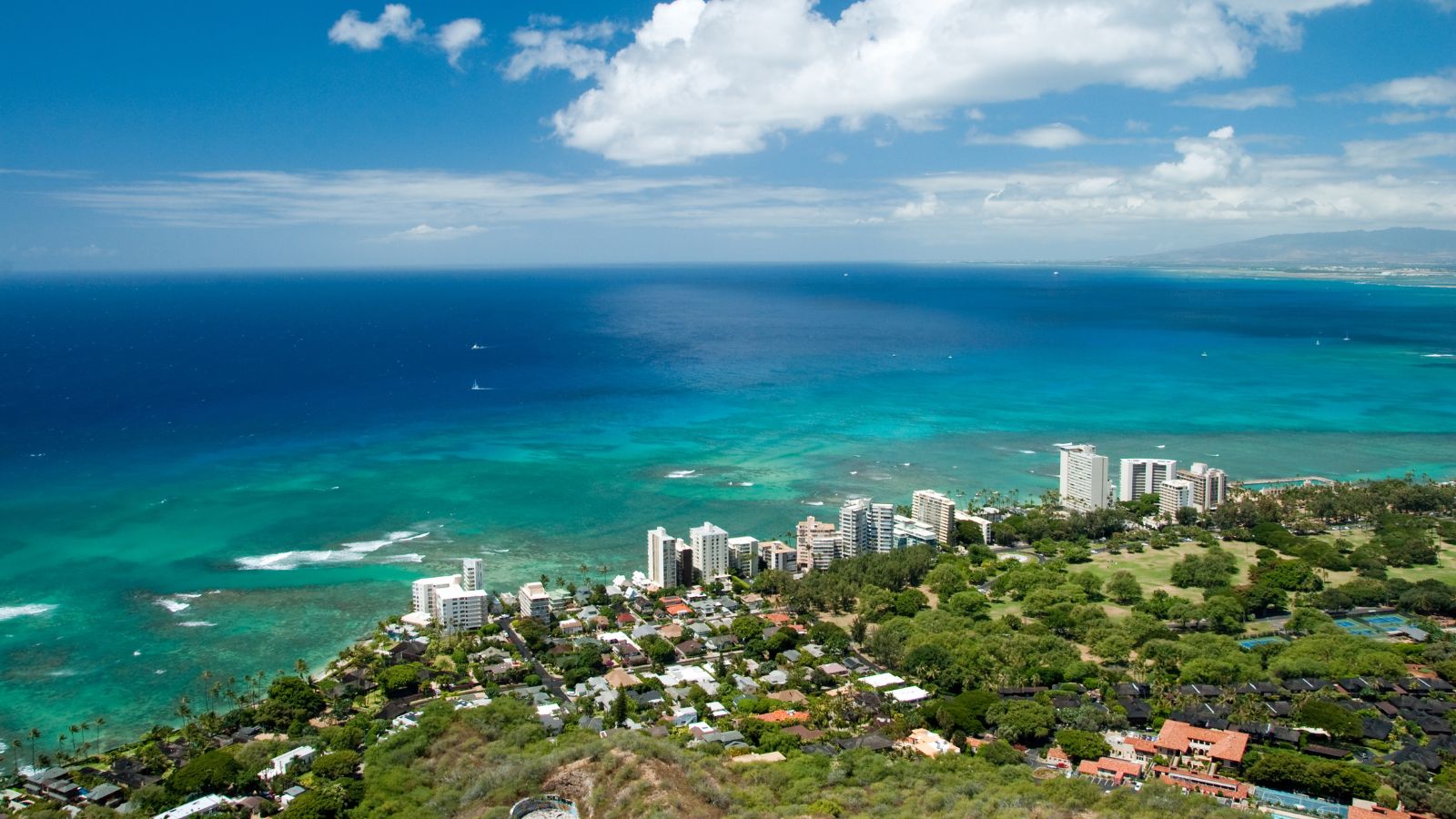 Aerial view of a coastal city with high-rises, green spaces, and turquoise ocean beneath a partly cloudy sky.