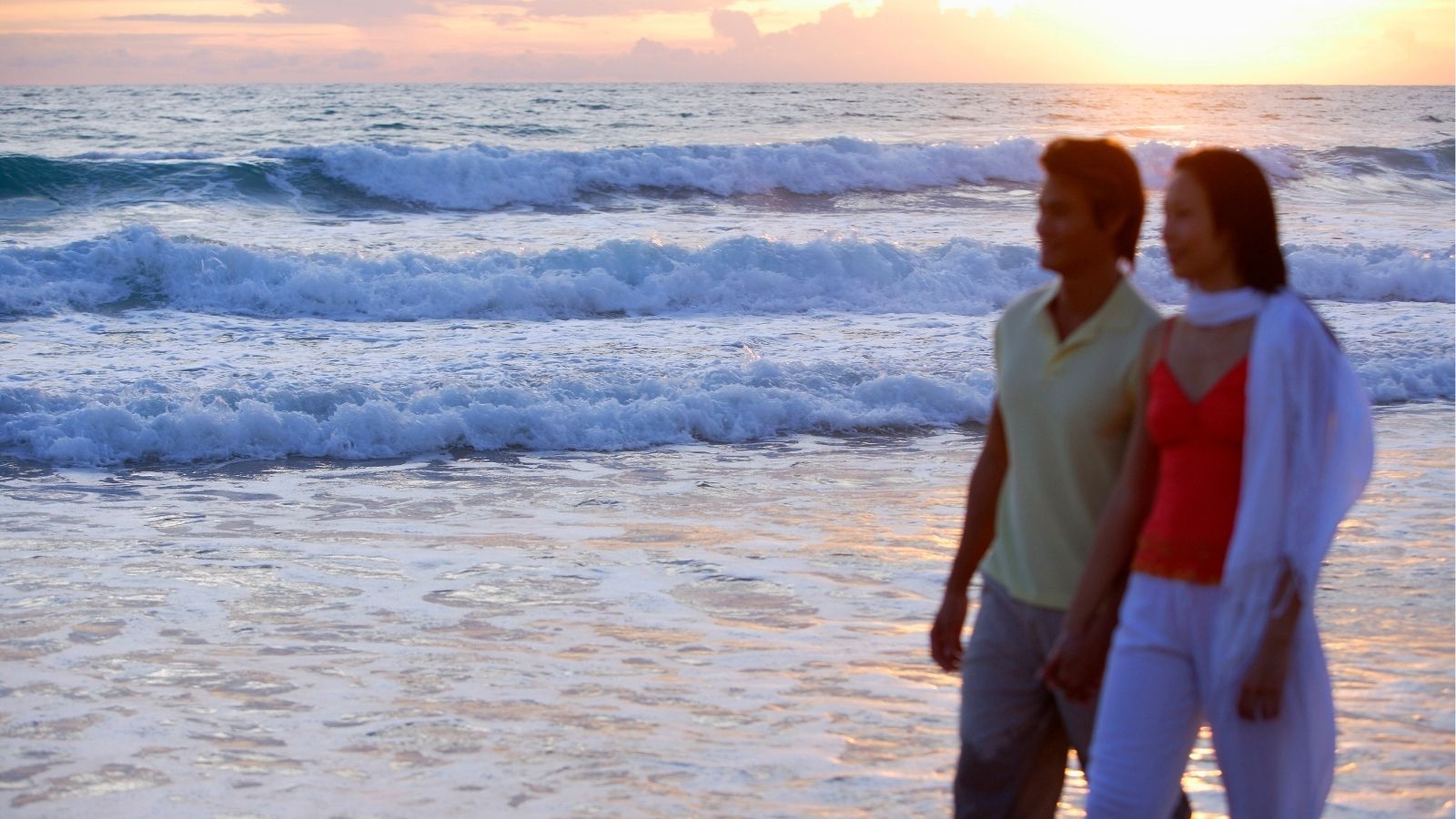 A couple walks on the shore at sunset, waves rolling in with the ocean behind them.