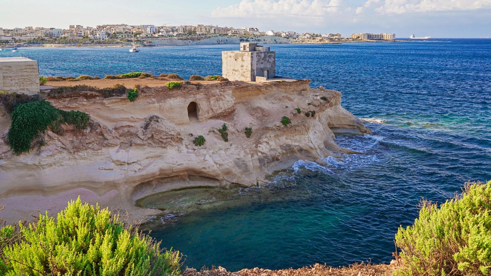 Stone structure on rocky cliff overlooks blue sea, with a distant town across the water beneath a partly cloudy sky.