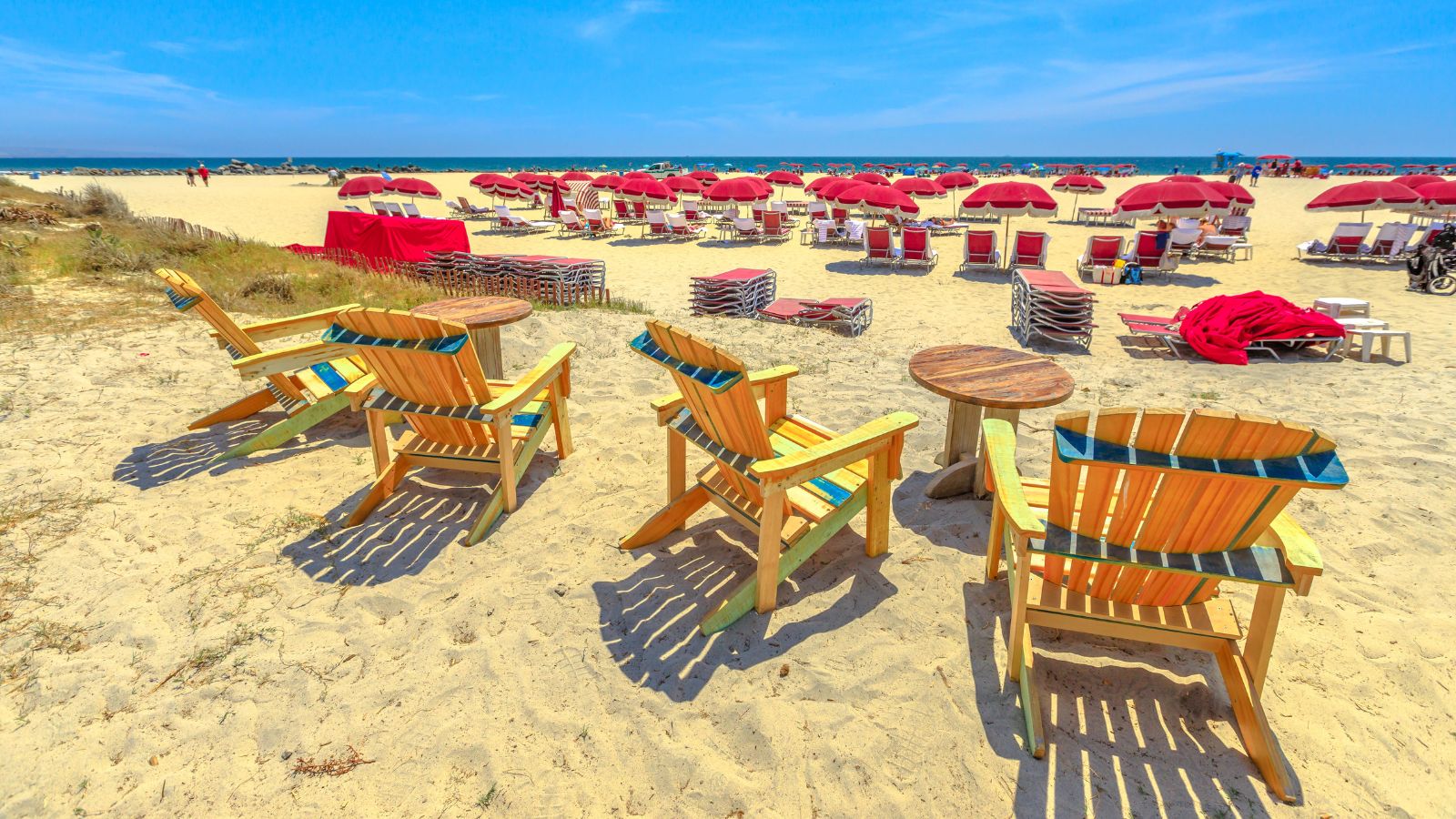 Wooden chairs overlook a sandy beach with red umbrellas, lounge chairs, blue sky, and the ocean in the background.