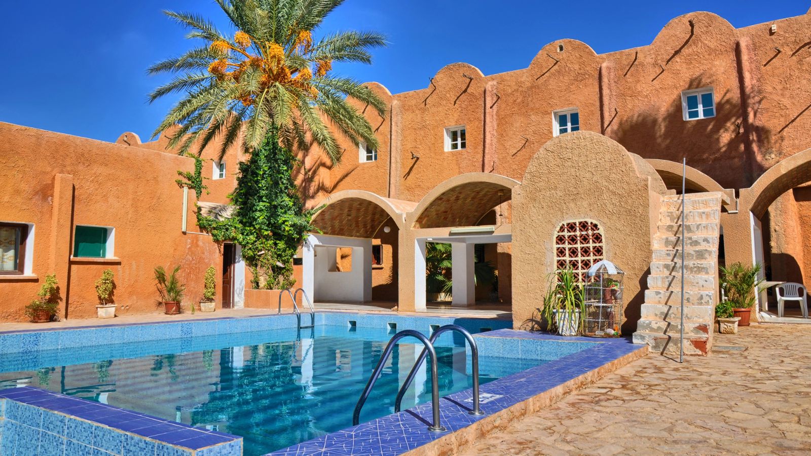 Rectangular blue-tiled pool framed by terra-cotta building, palm tree, potted plants, and white staircase beneath clear sky.