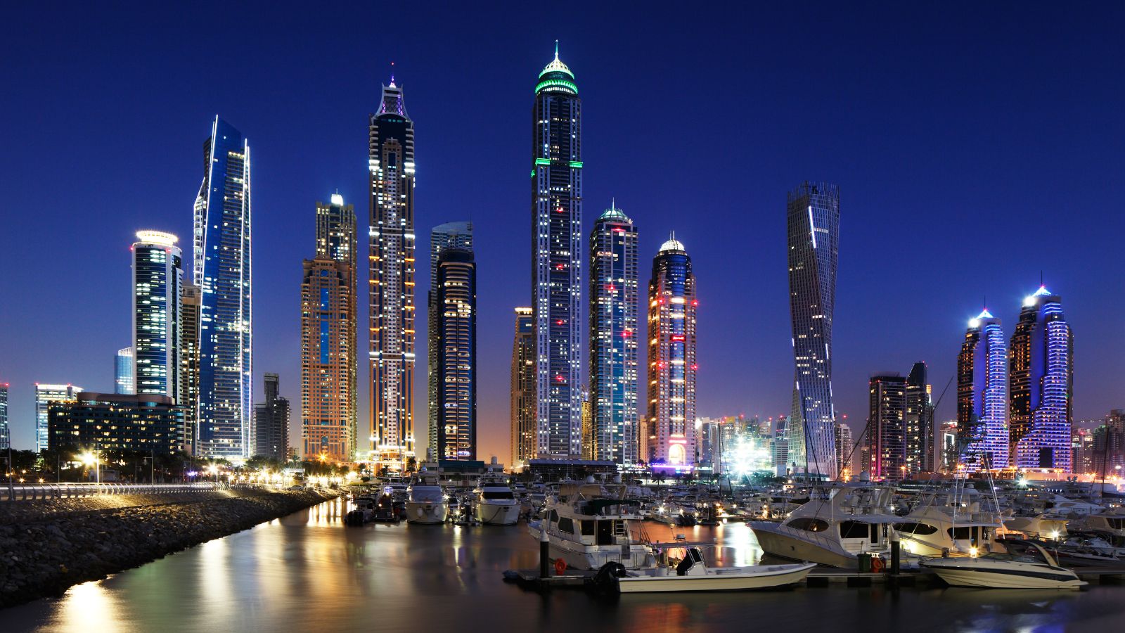 Illuminated skyscrapers and yachts reflect on marina waters beneath a clear dark blue night sky.