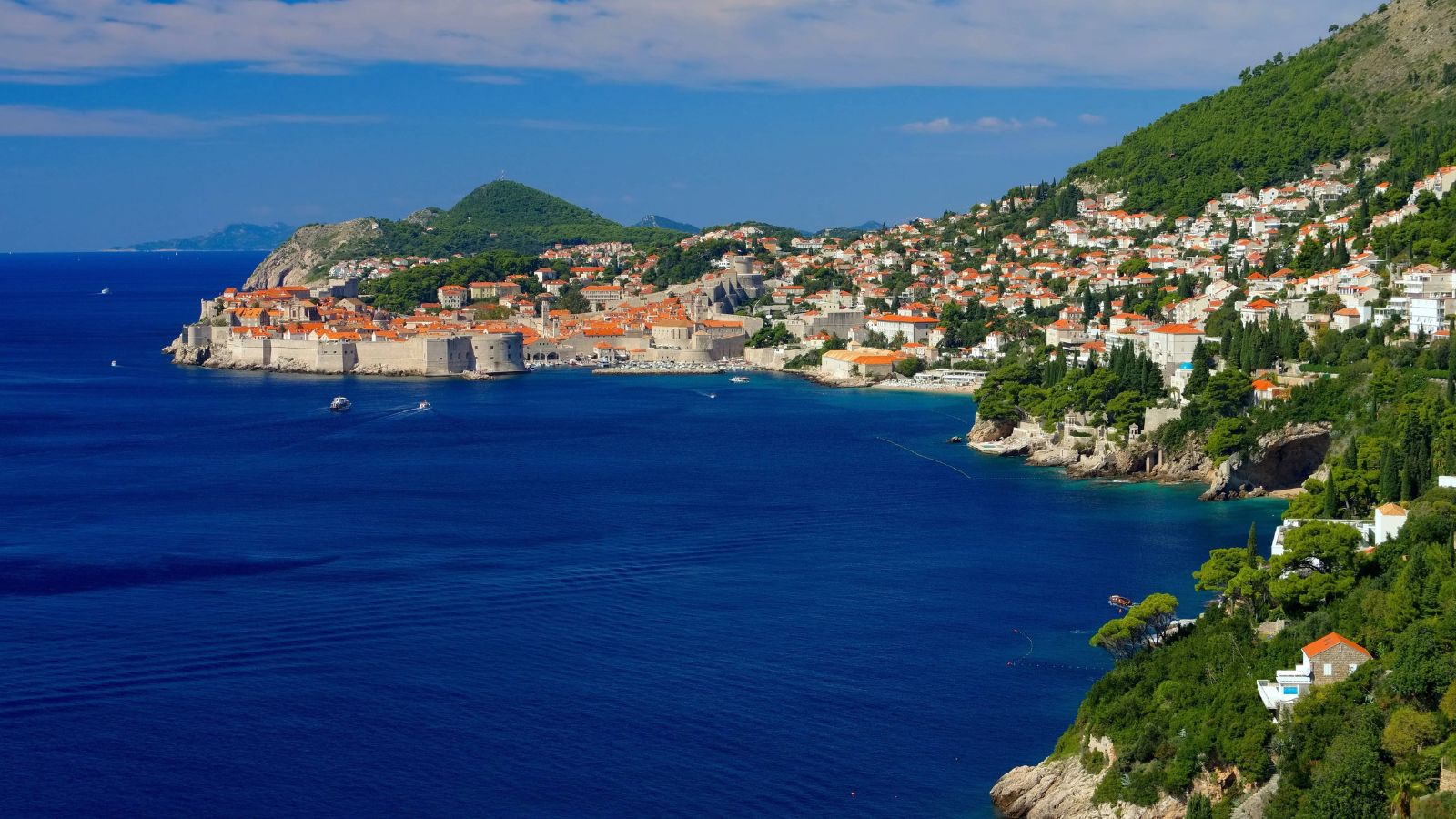 A coastal city with orange roofs and fortified walls sits by a deep blue sea, backed by green hills under a partly cloudy sky.