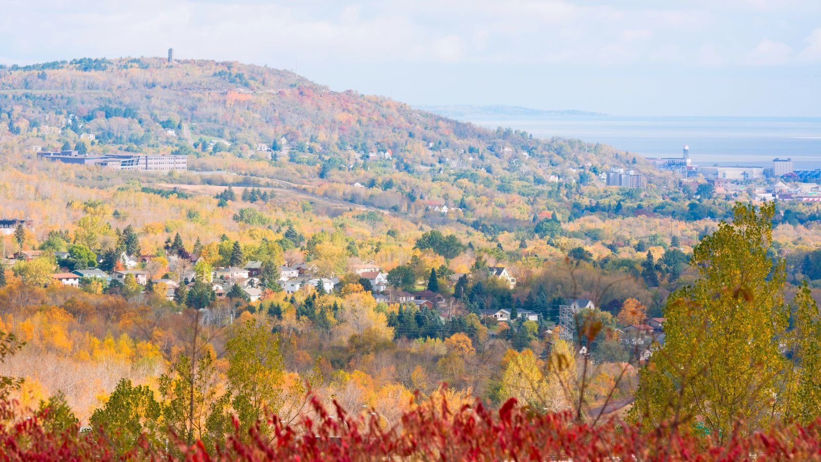 Hillside town with scattered houses and autumn trees overlooks a body of water beneath a partly cloudy sky.