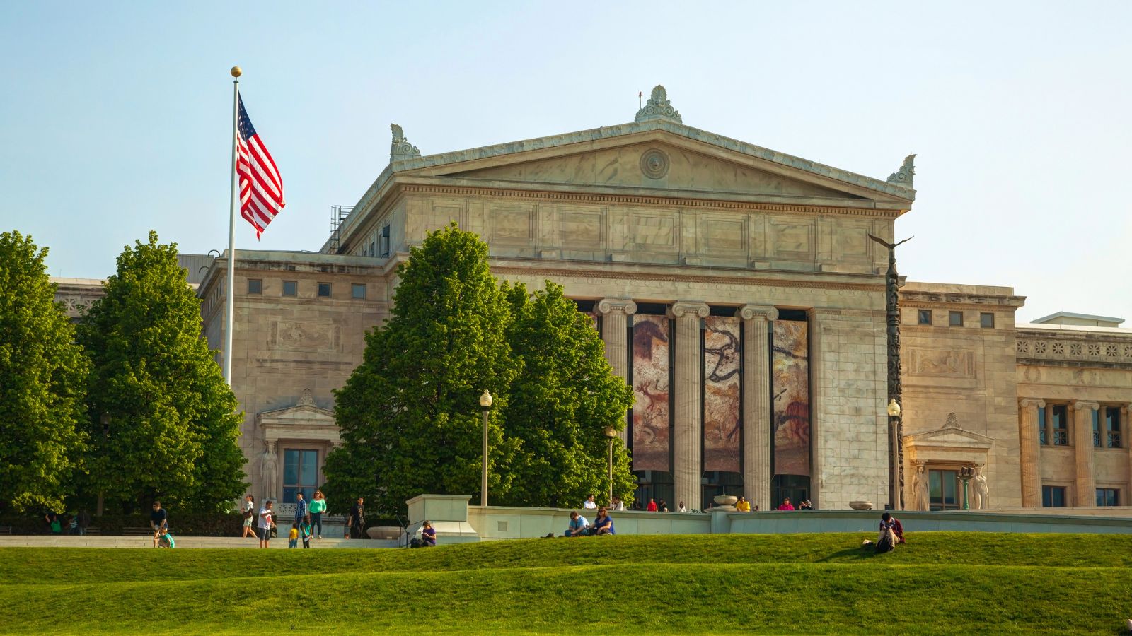 A neoclassical building with columns, banners, an American flag, trees, and people sitting on the grass in front.