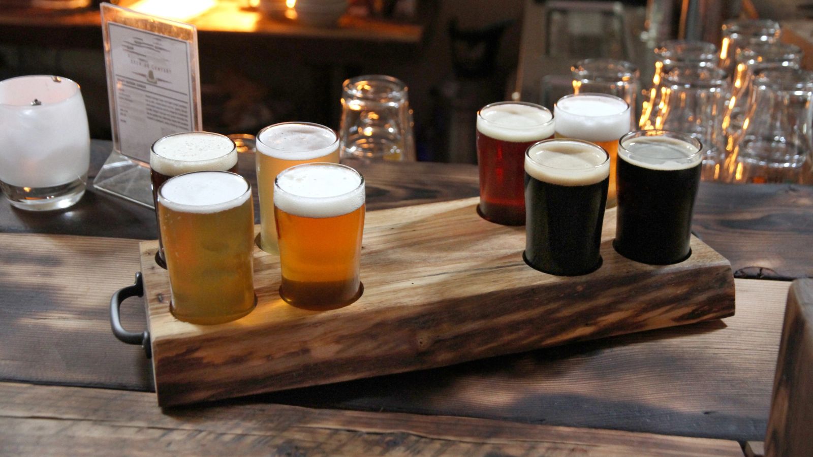 Eight small glasses of various colored beers sit on a wooden flight paddle atop a bar counter.