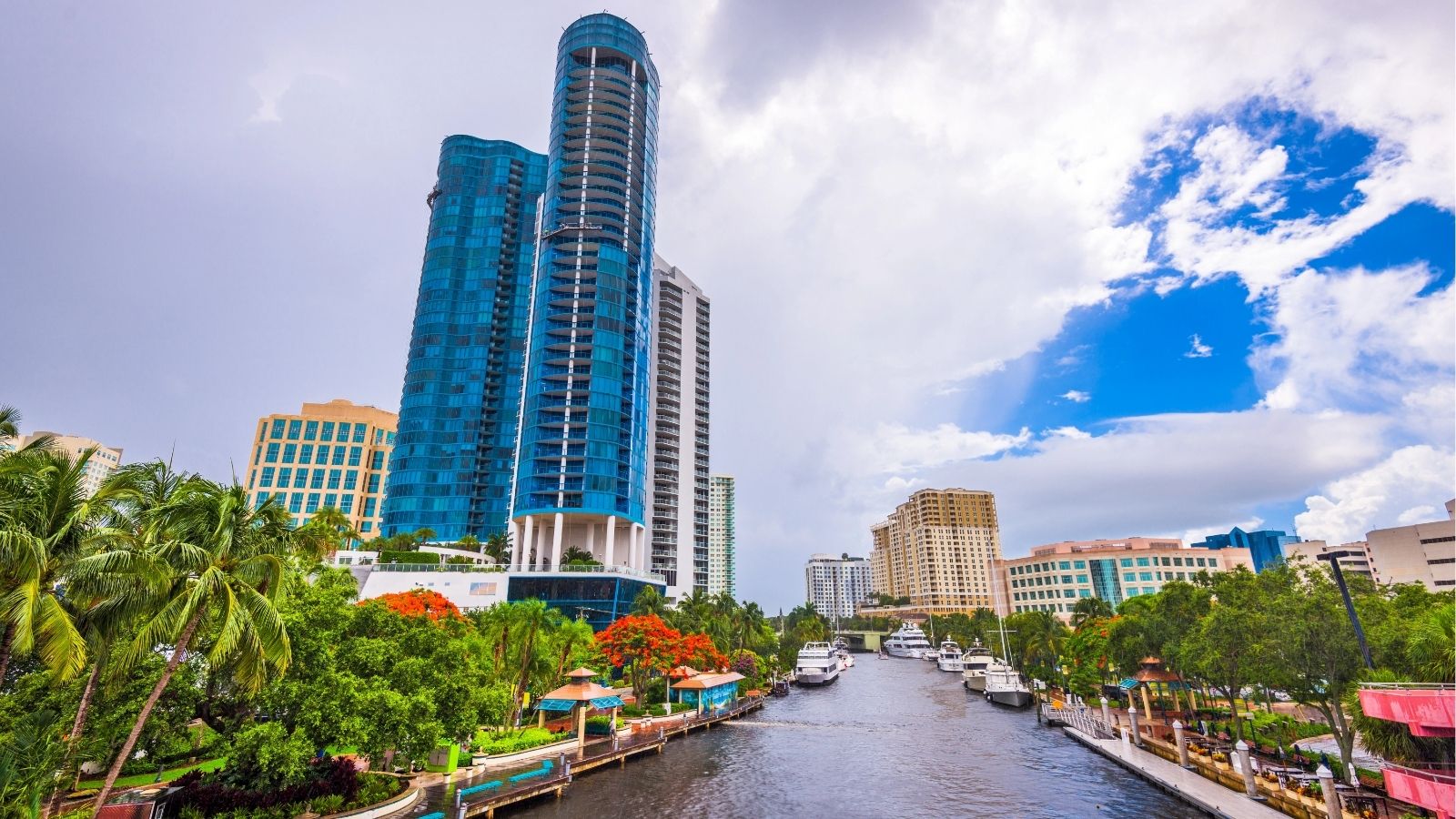 A blue high-rise and other modern buildings overlook a canal with boats, palm trees, and greenery beneath a partly cloudy sky.