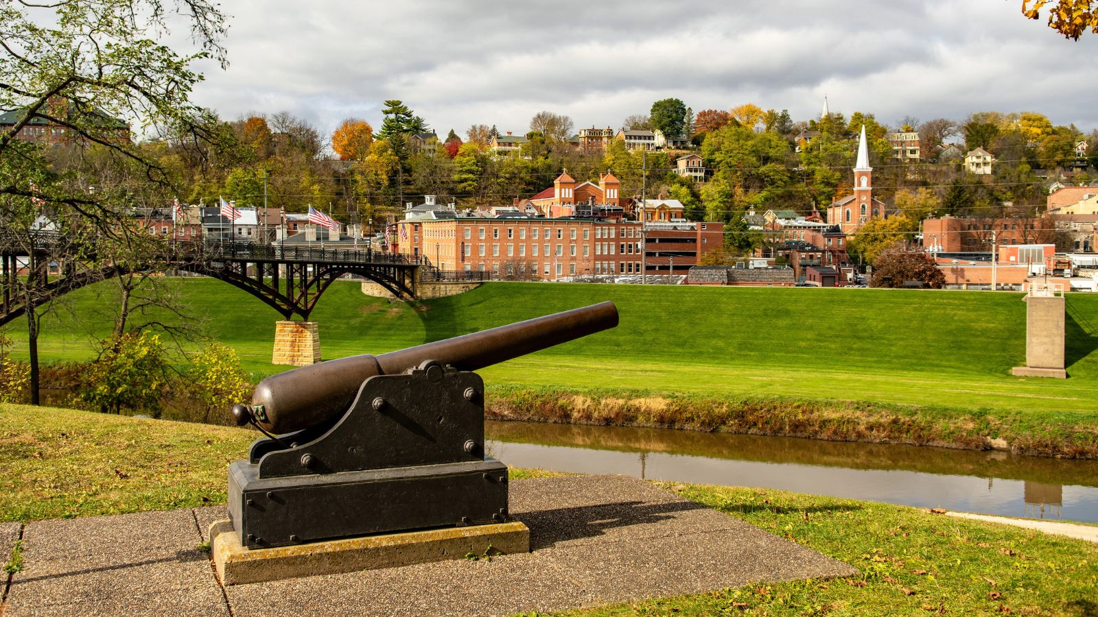 A historic cannon faces a grassy area, river, and town with brick buildings, church, and autumn trees in the background.