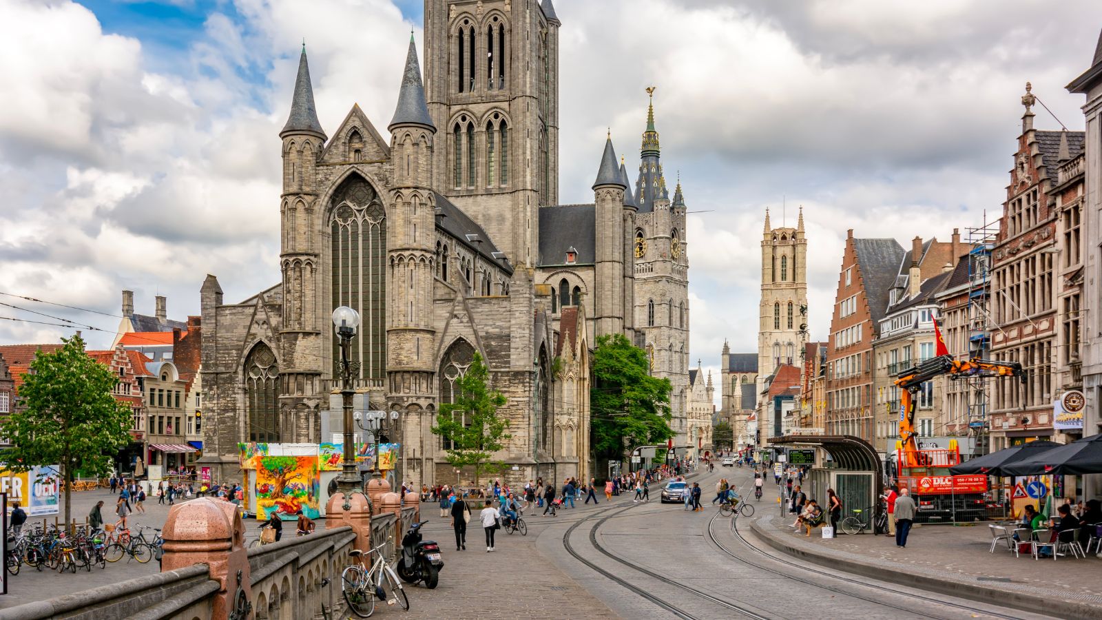 St. Nicholas' Church in Ghent rises above tram tracks, cyclists, pedestrians, and outdoor cafes on a partly cloudy day.