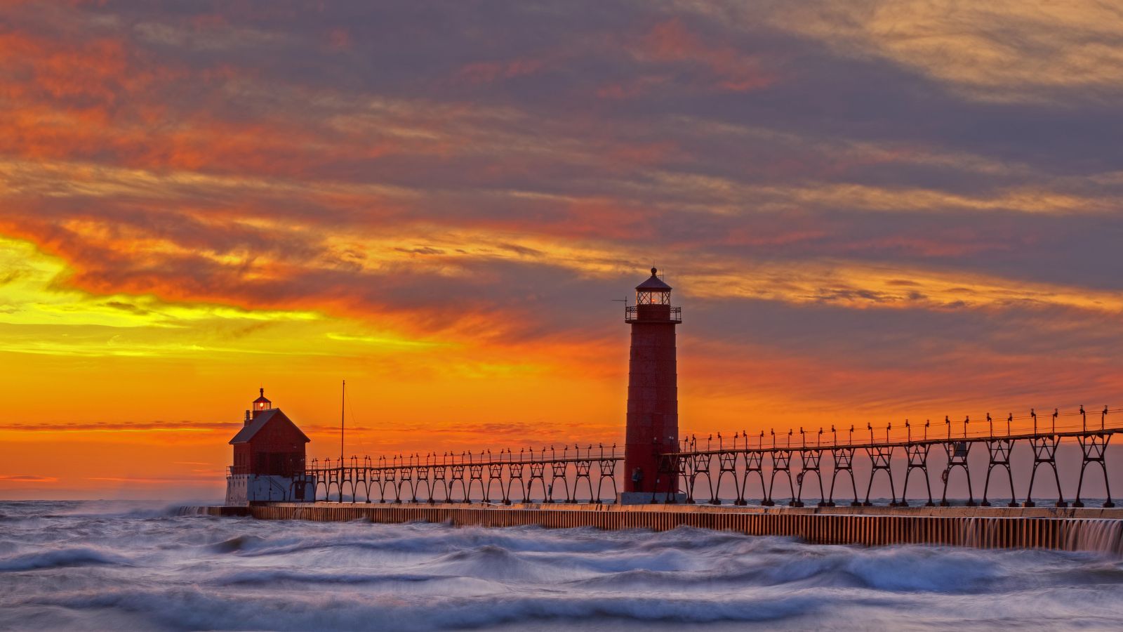 A lighthouse and pier jut into rough waters at sunset beneath an orange-yellow sky with scattered clouds.