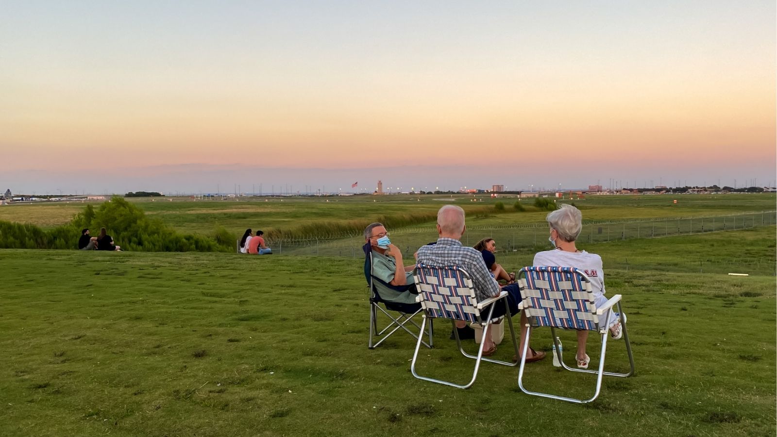 Three people on lawn chairs face a sunset over a grassy field, with others farther back and buildings on the distant horizon.