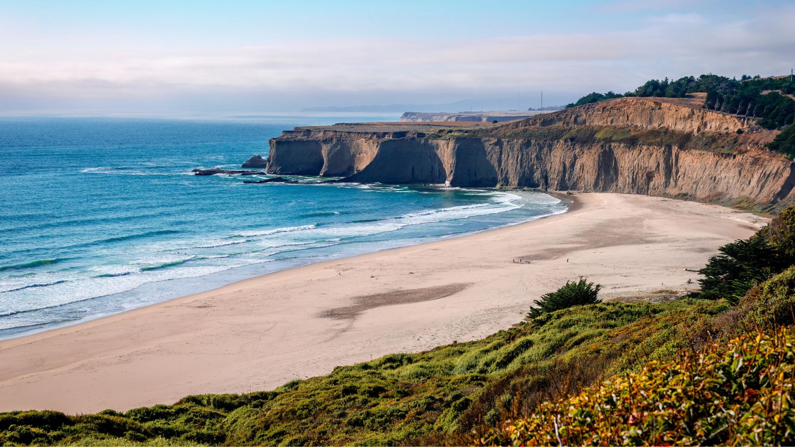 Wide sandy beach with gentle waves, flanked by tall cliffs and greenery beneath a partly cloudy sky.